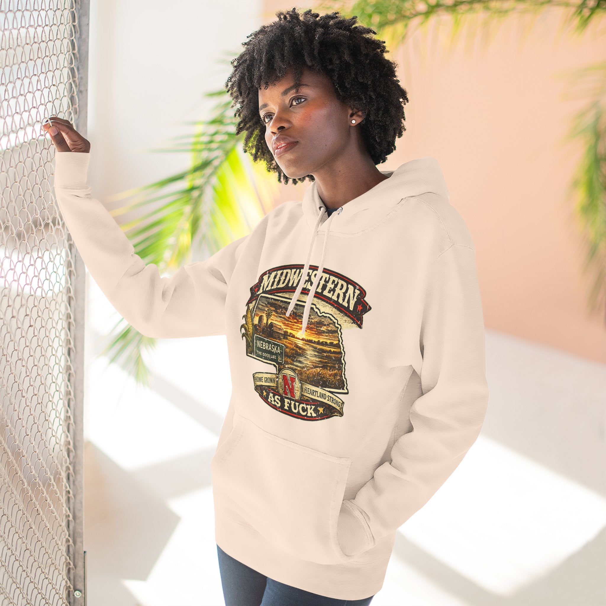 Woman in the pale pink Nebraska As Fuck hoodie leaning against a fence outdoors