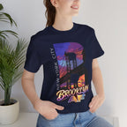 Navy t-shirt worn by a model, close-up of chest print with 'B' Brooklyn Bridge sunset skyline graphic, plant in background
