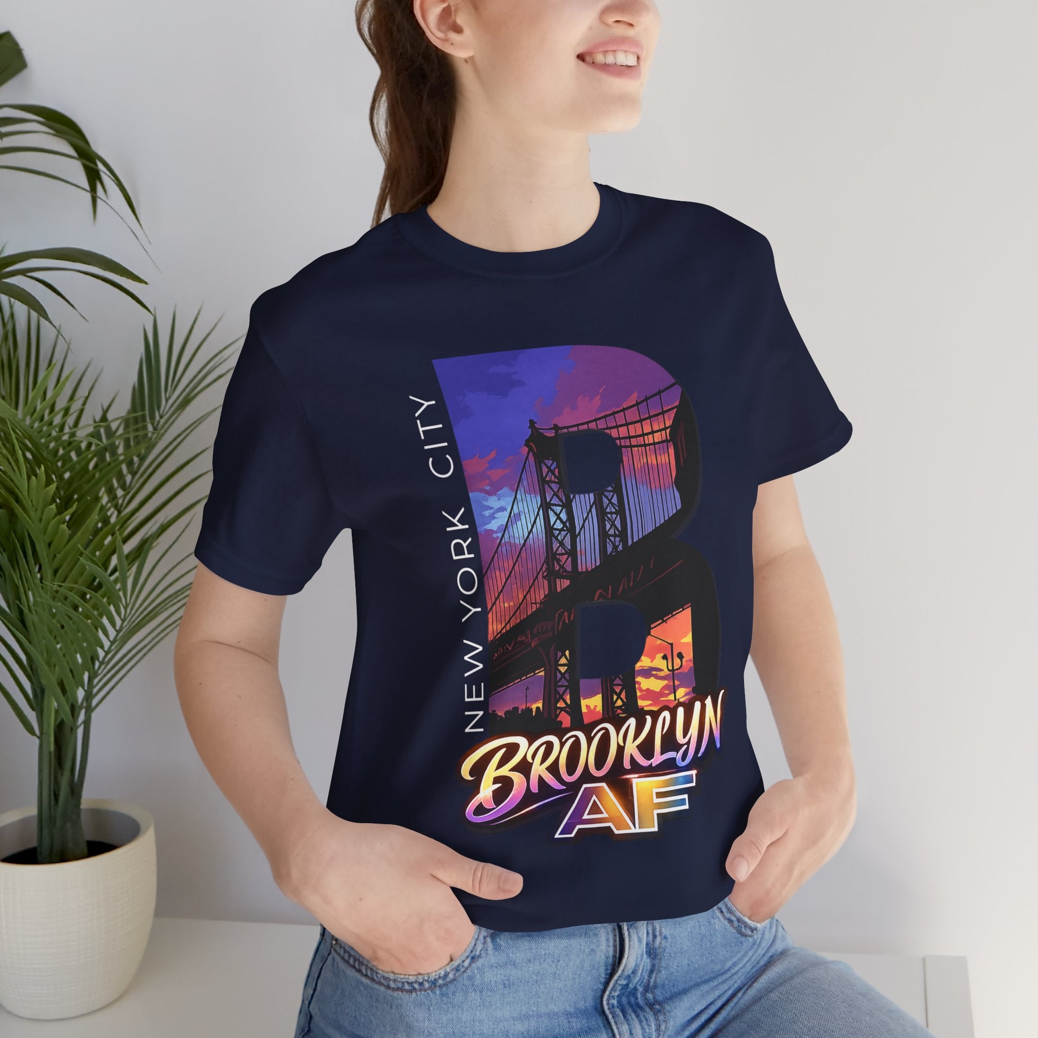Navy t-shirt worn by a model, close-up of chest print with 'B' Brooklyn Bridge sunset skyline graphic, plant in background