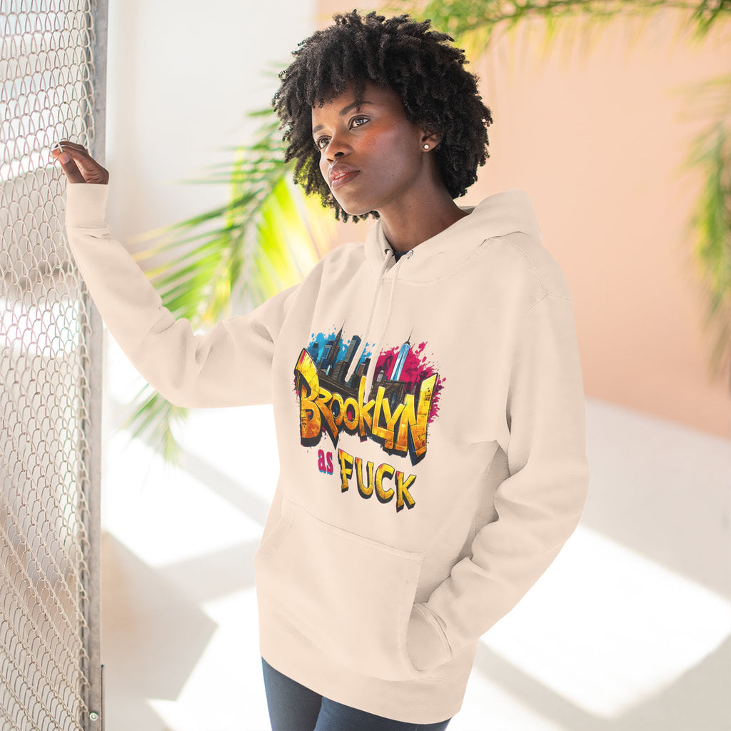 Oatmeal heather hoodie worn by a female model near a wire fence, front view, graffiti Brooklyn As Fuck NYC skyline print.