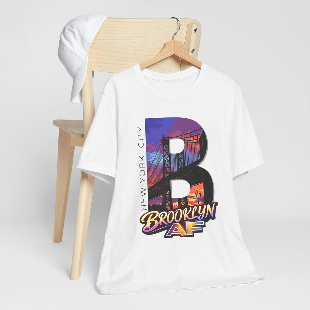 White tee on a wooden chair with a white cap, front view, showing the "B" Brooklyn Bridge sunset skyline graphic.