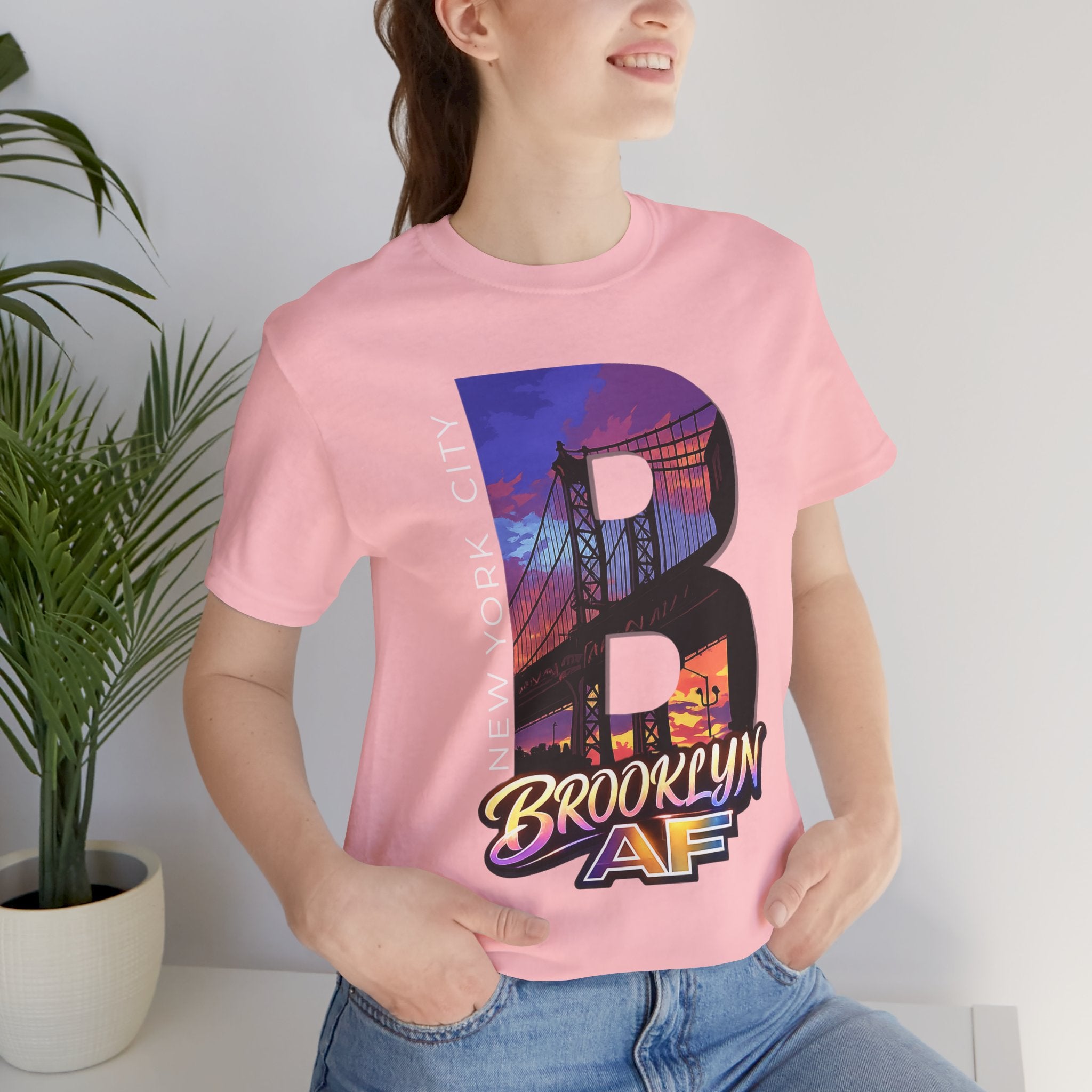Pink t-shirt worn by a model, close-up of chest print with 'B' Brooklyn Bridge sunset skyline graphic, plant in background