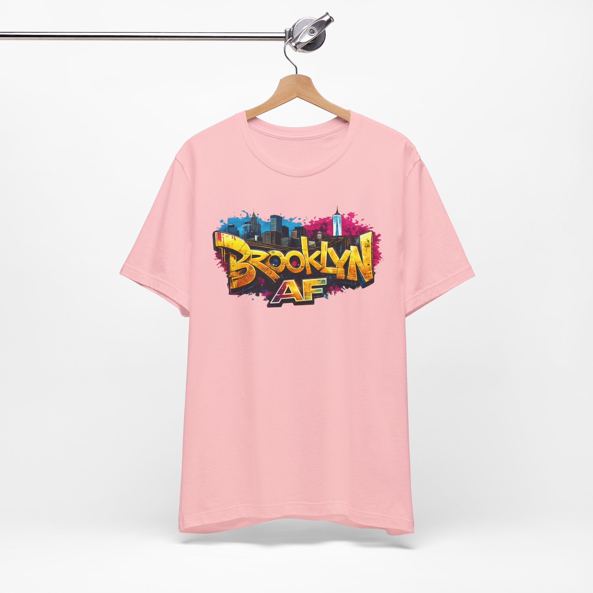 Pink t-shirt on a wooden hanger, front view, showing graffiti-style Brooklyn AF lettering over a skyline paint splash.