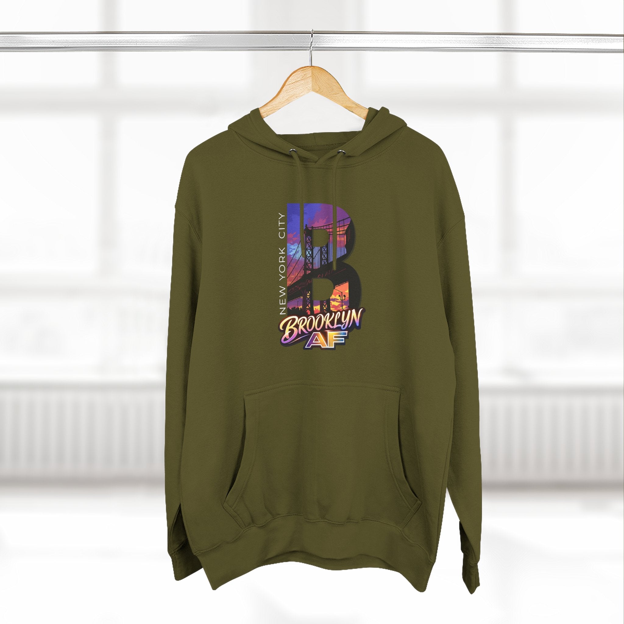 Army green hoodie on a wooden hanger against a blurred window, showing the oversized "B" skyline graphic.
