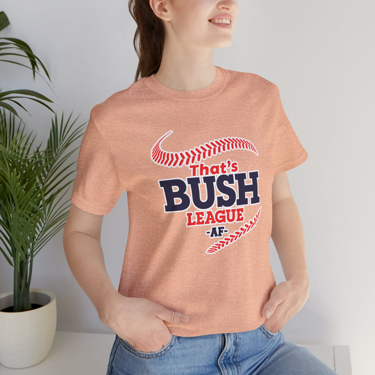 Woman wearing the Heather Peach Bush League AF tee beside a plant, navy lettering visible