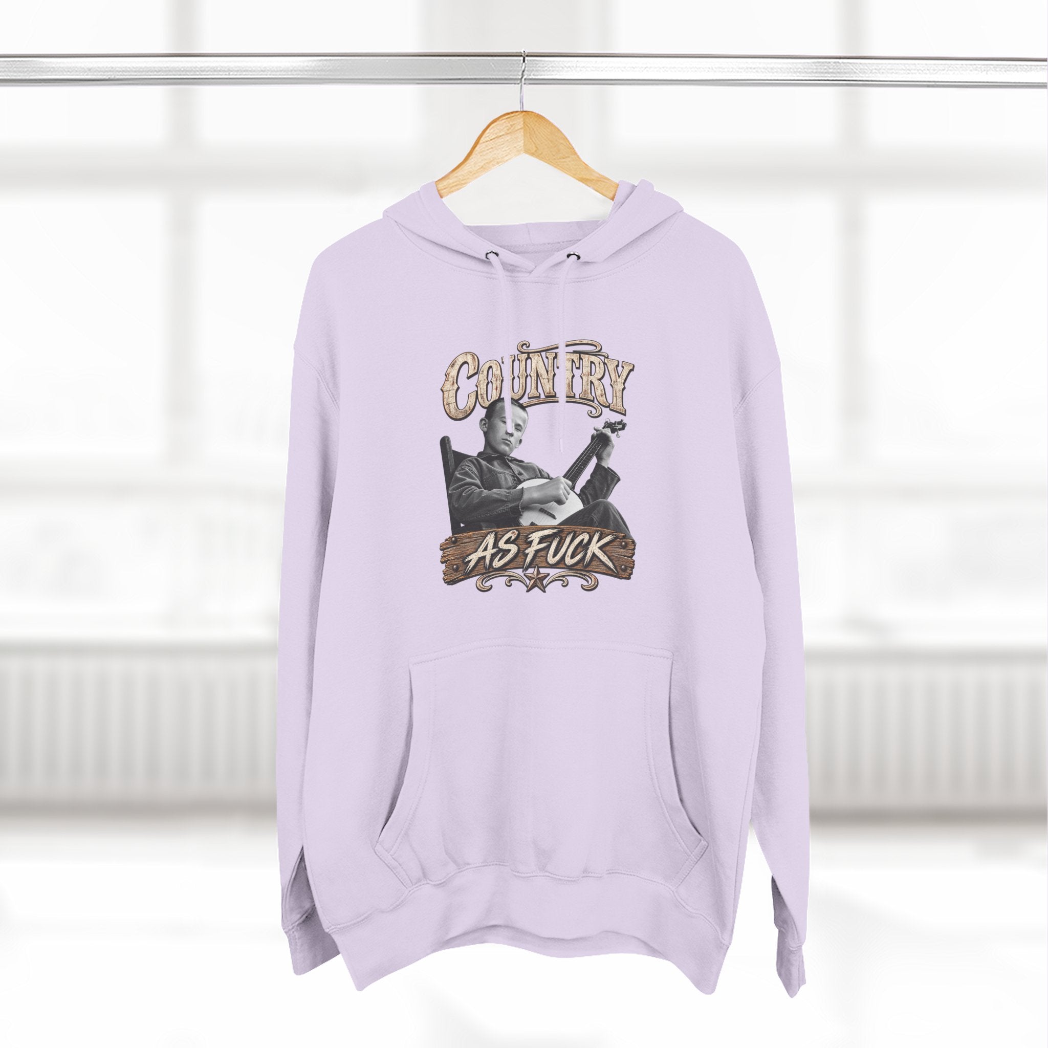Lilac Country As Fuck hoodie hanging on a wooden hanger by a window