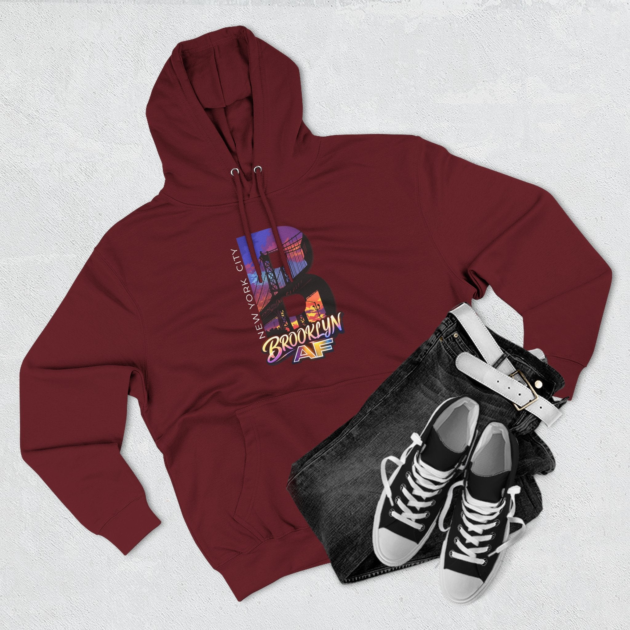 Red hoodie flat lay styled with jeans, white belt, and sneakers, showing the oversized "B" Brooklyn Bridge skyline graphic.
