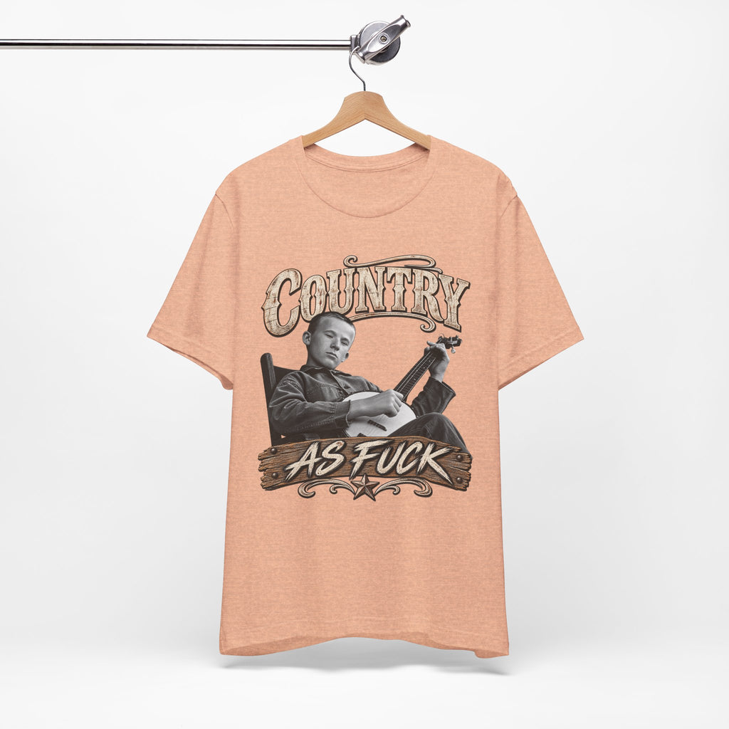 Heather Peach Country As Fuck tee on a wooden hanger against a white wall