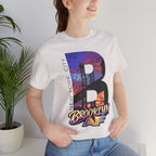 White tee worn by a female model near a plant, close-up of the "B" Brooklyn Bridge sunset skyline chest print.