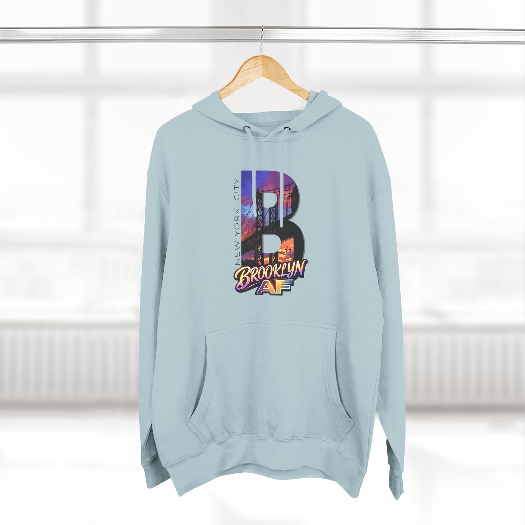 Pale blue hoodie hanging on a wooden hanger, bold B with a Brooklyn Bridge sunset skyline graphic.