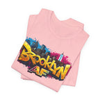 Pink t-shirts folded in a stack, featuring graffiti Brooklyn AF lettering over a skyline paint-splash design.