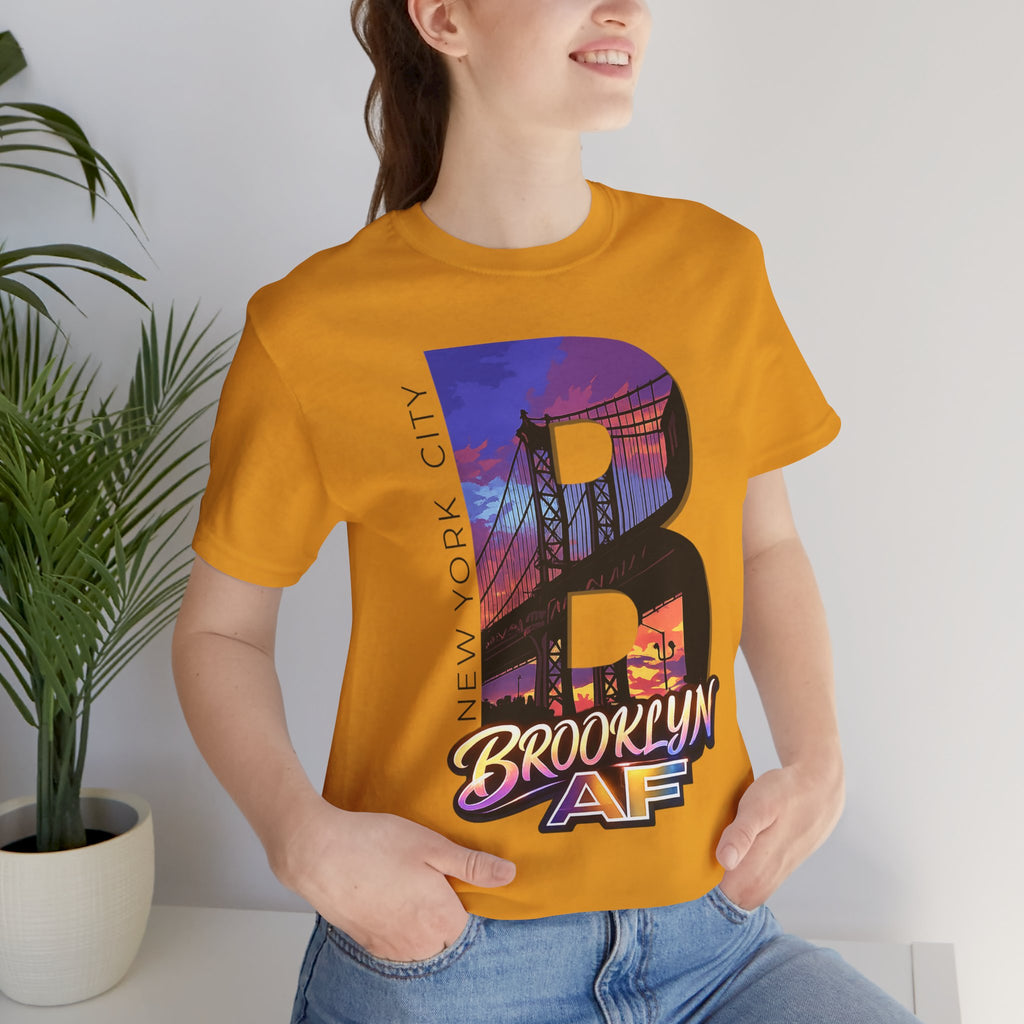 Mustard tee on a female model, close-up of the "B" Brooklyn Bridge sunset skyline chest print near a potted plant.