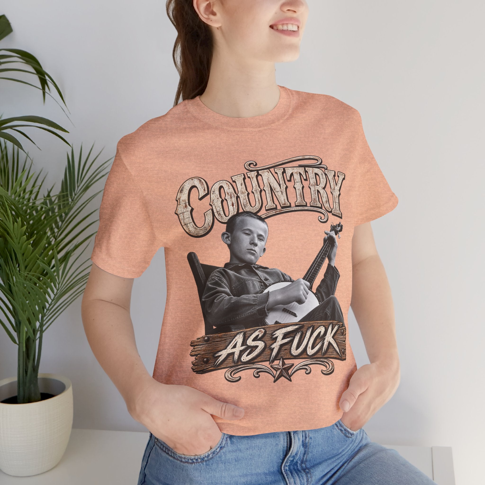 Woman beside a plant in the Heather Peach Country As Fuck tee
