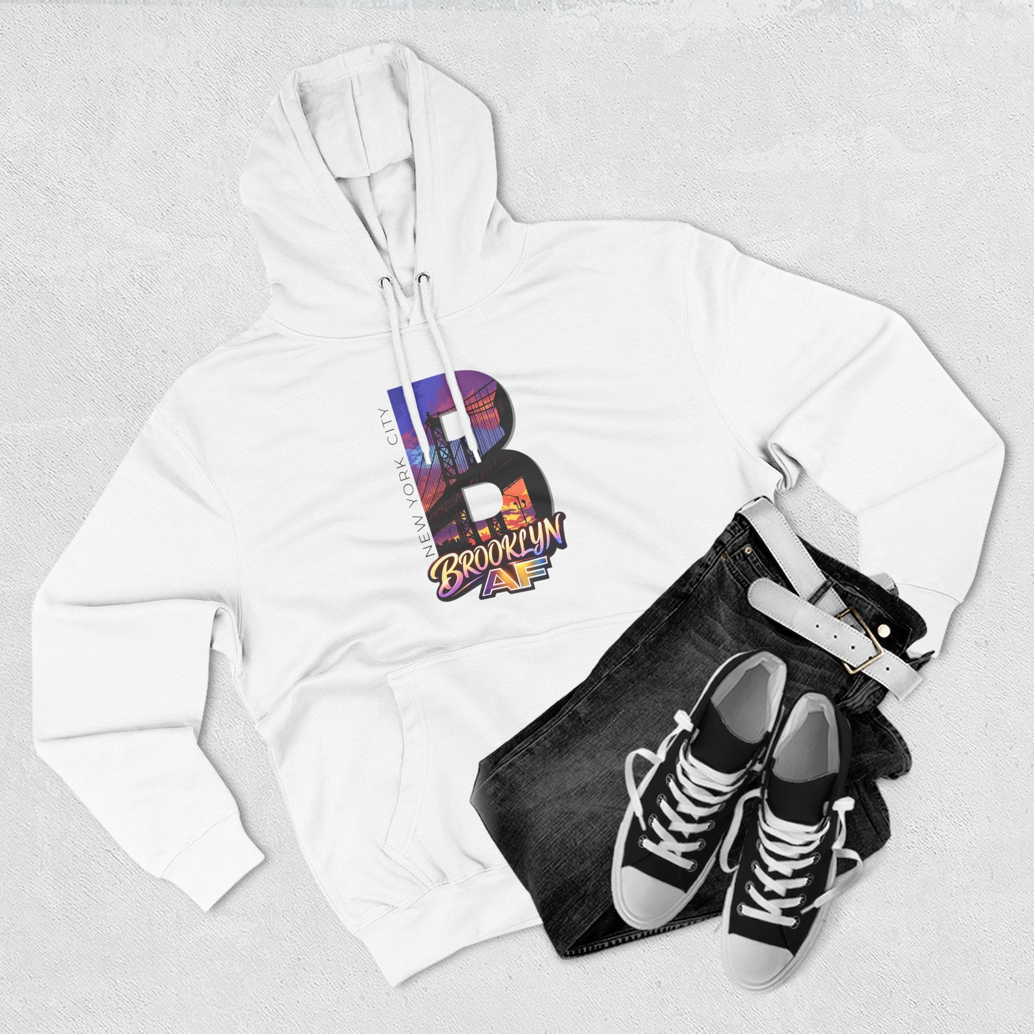 White hoodie styled with black jeans, belt, and sneakers, bold B showing a Brooklyn Bridge sunset skyline graphic.