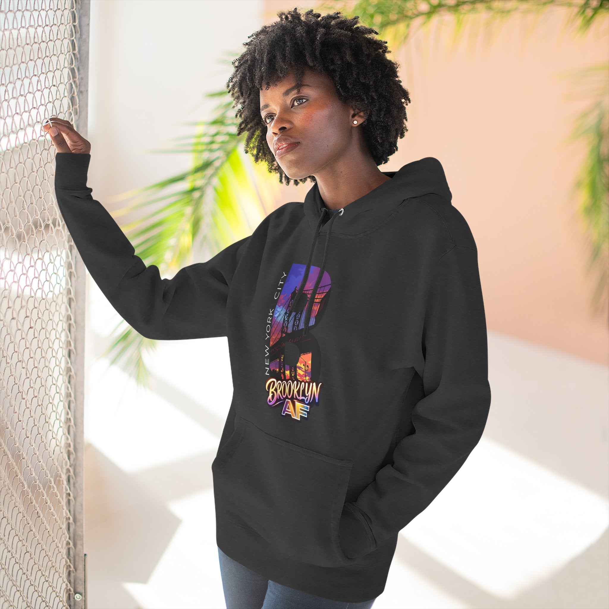 Black hoodie worn by a female model outdoors, side view with hand on a fence, oversized "B" skyline graphic on chest.