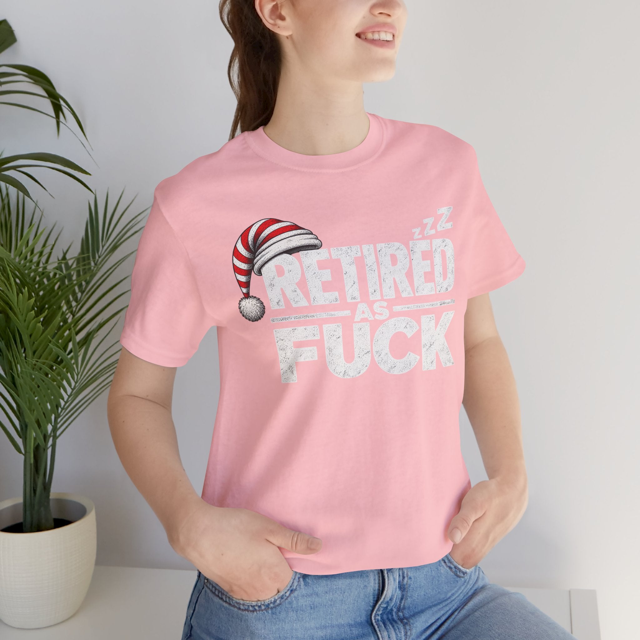 Woman in the Pink Retired As Fuck tee and jeans standing beside a houseplant
