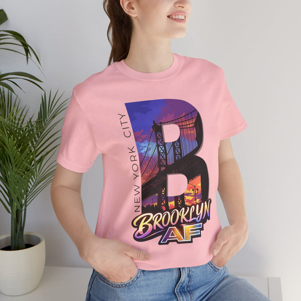 Pink tee worn by a female model near a plant, close-up of the "B" Brooklyn Bridge sunset skyline chest print.