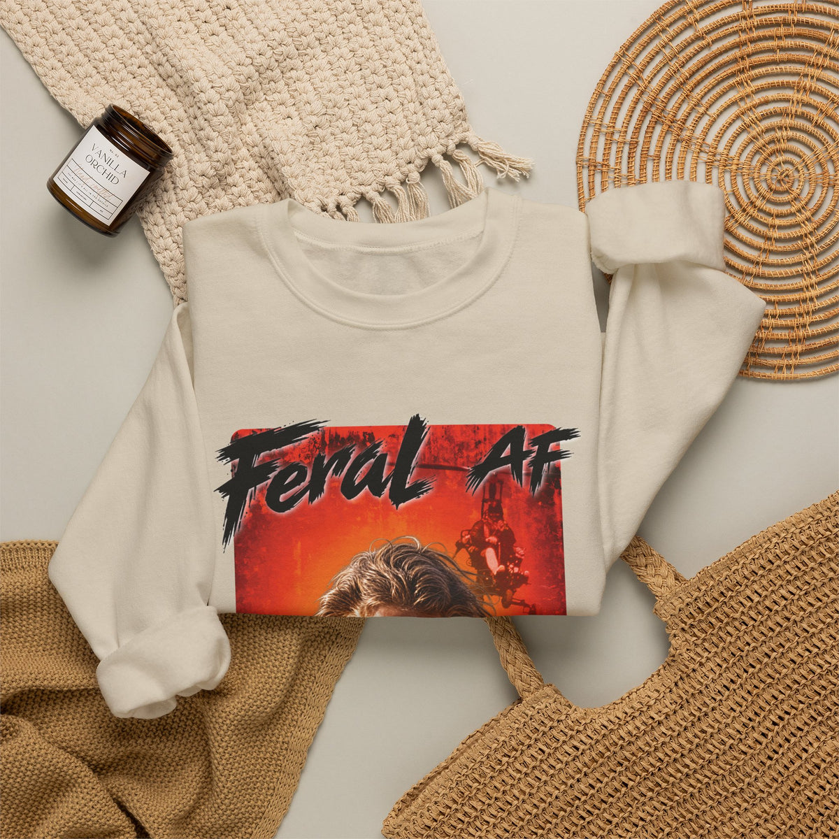 Sand Feral AF sweatshirt folded on a woven mat with a knit throw and a candle