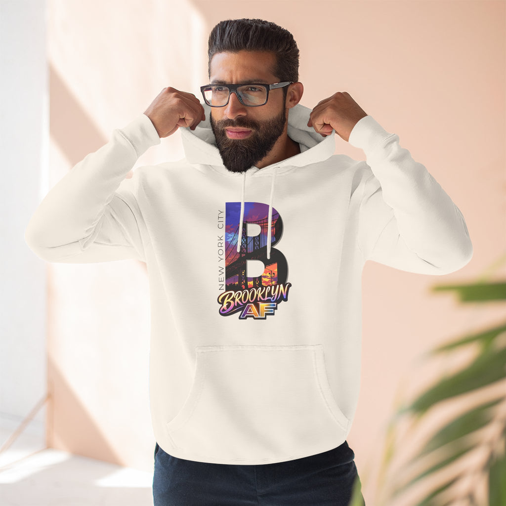 White hoodie worn by a bearded male model pulling up the hood, bold B with a Brooklyn Bridge sunset skyline graphic.