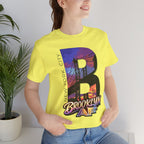Yellow t-shirt worn by a model, close-up of chest print showing the 'B' with Brooklyn Bridge sunset skyline graphic