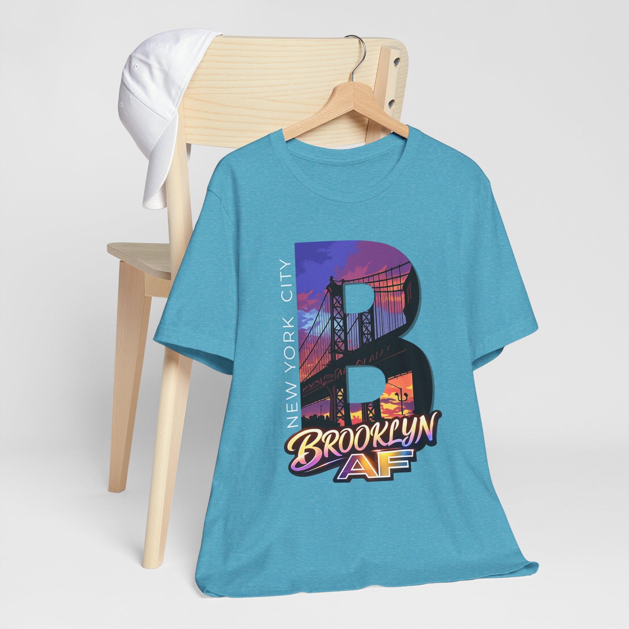 Heather aqua t-shirt hanging on a wooden hanger, front view, with a bold B and purple-orange Brooklyn Bridge sunset graphic.