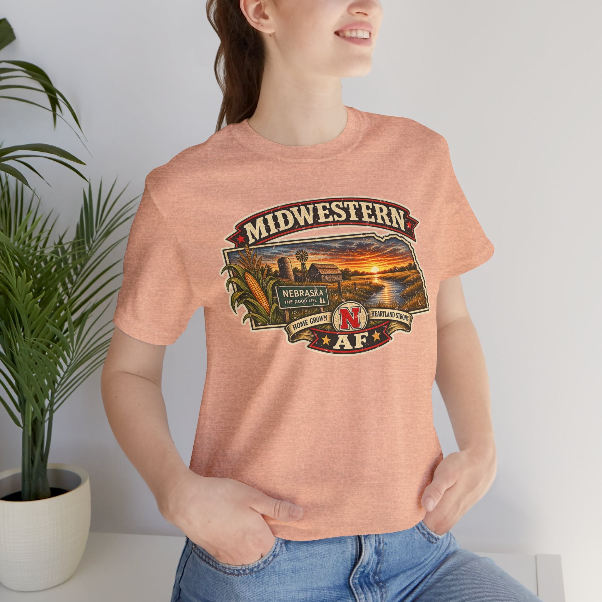 Heather peach Nebraska tee worn indoors, close on the Midwestern banner over a Nebraska-shaped farm scene