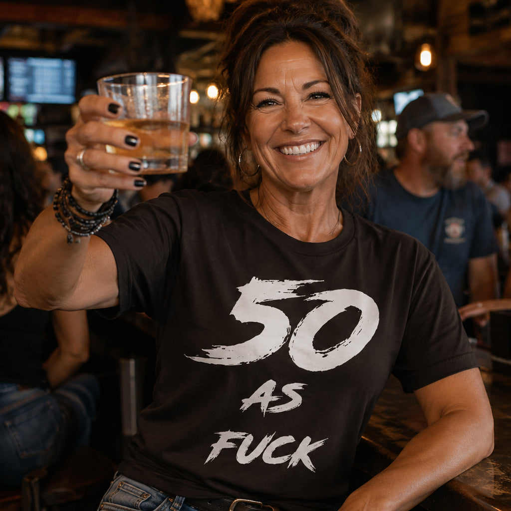 Woman in a bar raising a drink, wearing the dark brown 50 AS FUCK birthday tee