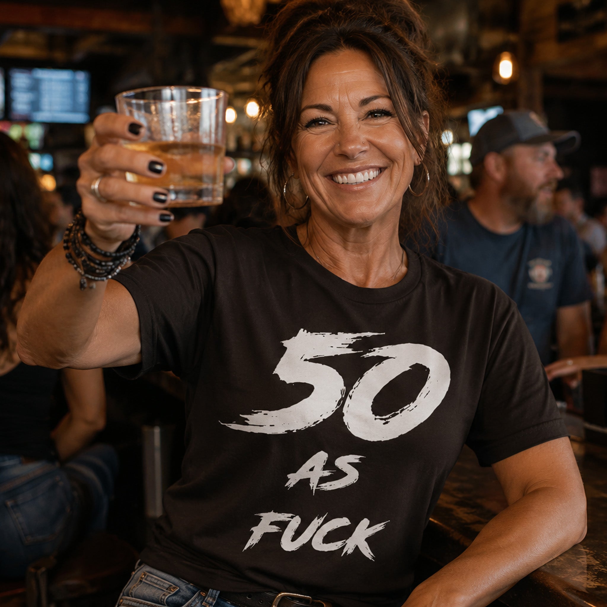 Woman in a bar raising a drink, wearing the dark brown 50 AS FUCK birthday tee