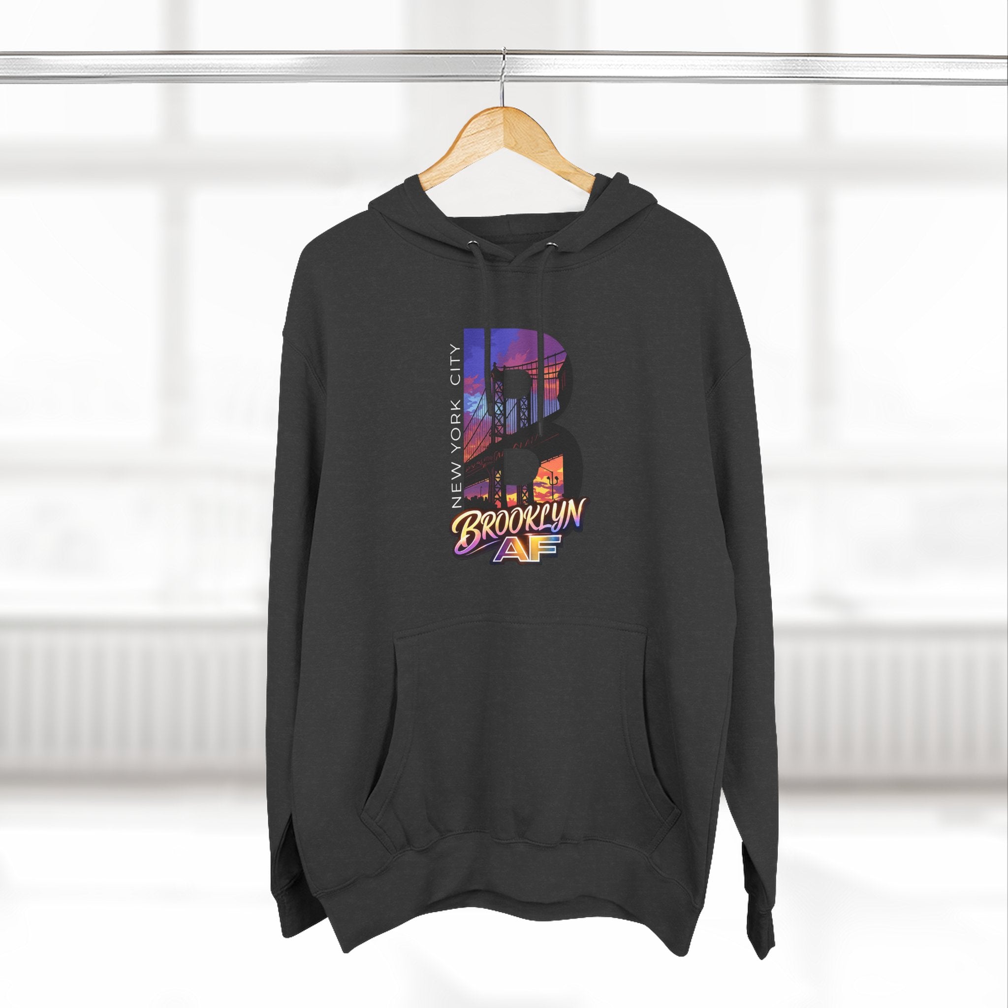 Black hoodie on a wooden hanger against a blurred window, showing the oversized "B" Brooklyn Bridge skyline graphic.
