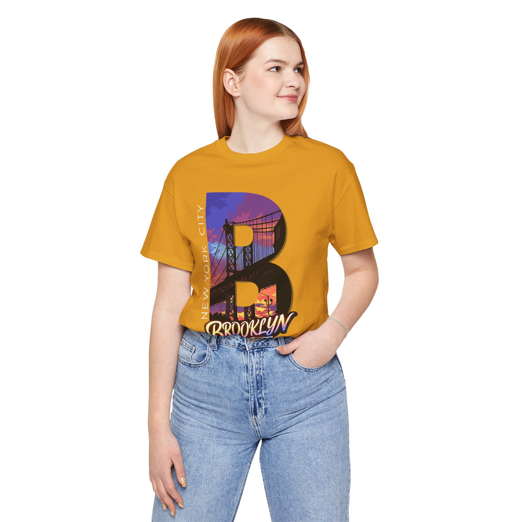 Mustard yellow t-shirt worn by a female model, front view, with a bold B and purple-orange Brooklyn Bridge sunset graphic.