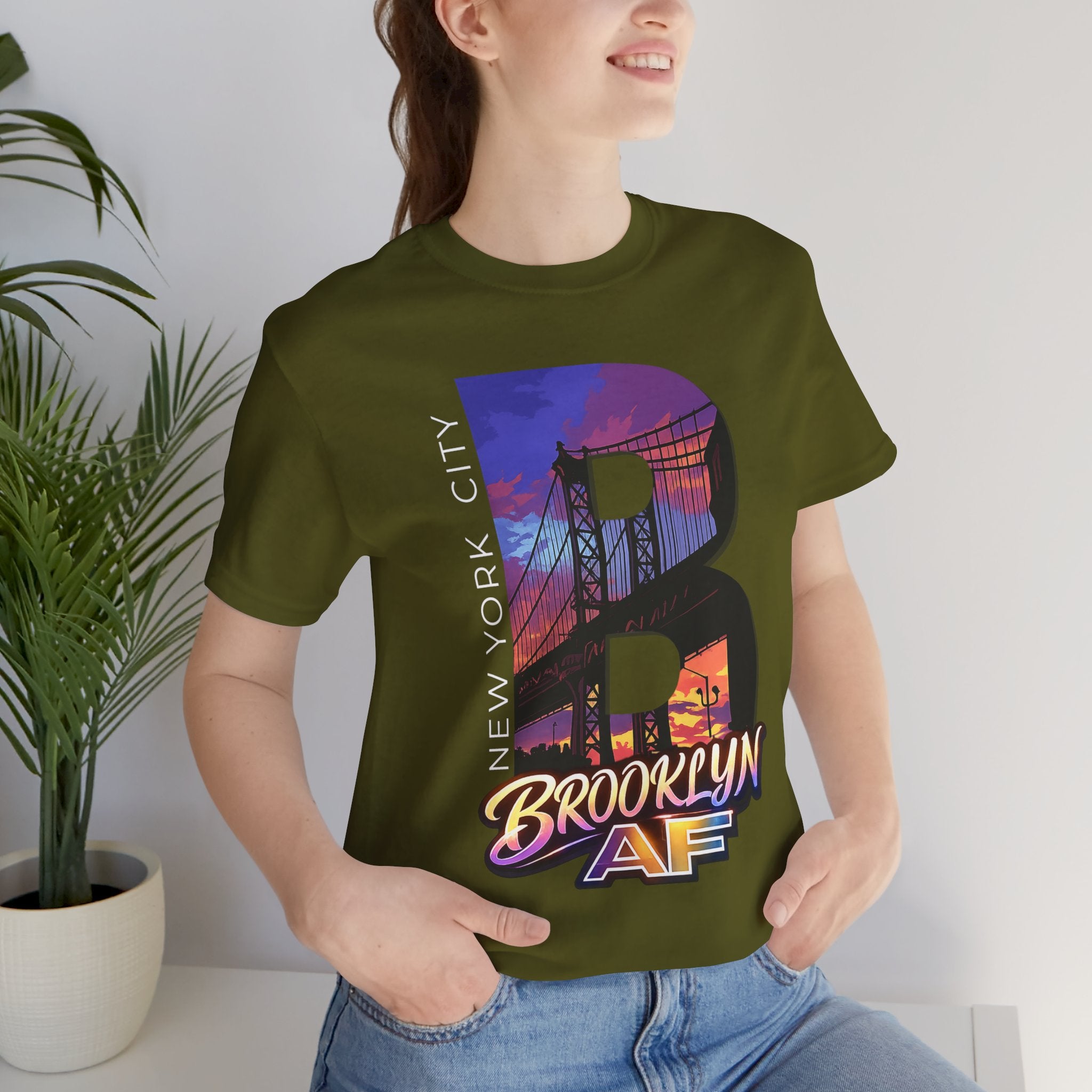 Olive green t-shirt worn by a model, close-up of chest print with 'B' Brooklyn Bridge skyline graphic, plant in background