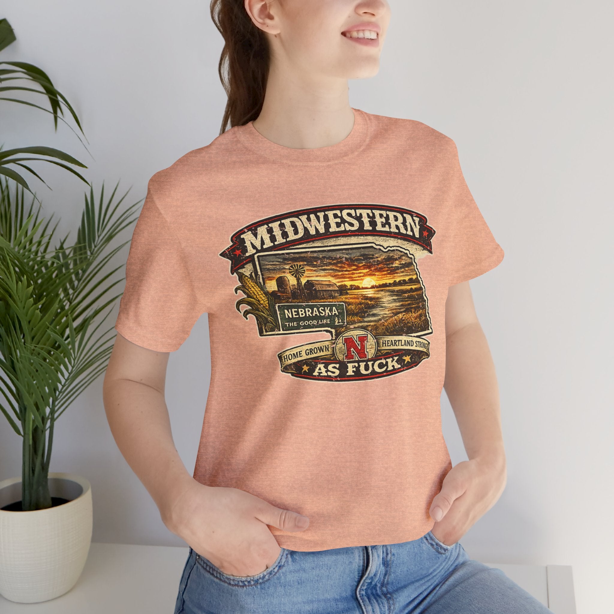Woman in the heather peach Nebraska As Fuck tee indoors beside a potted plant