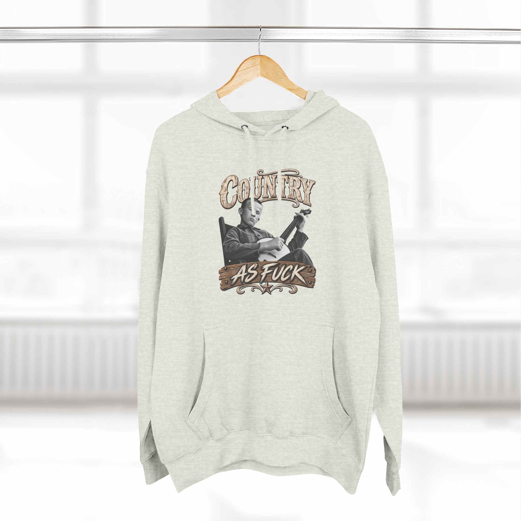 Oatmeal Heather Country As Fuck hoodie on a wooden hanger by a window