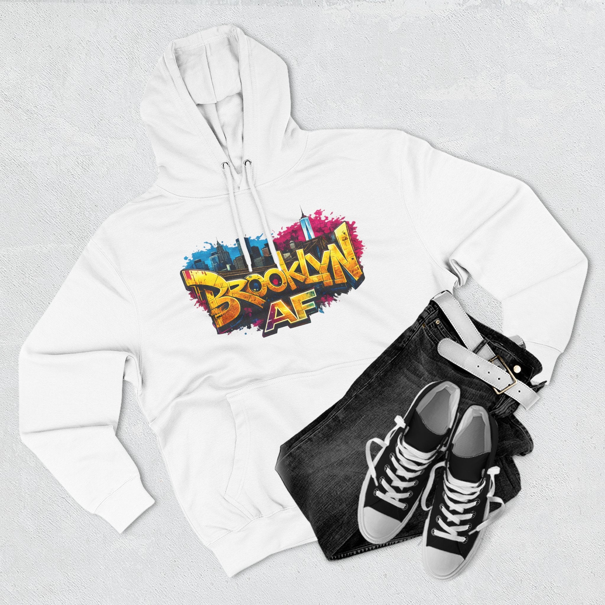 White hoodie flat lay styled with black jeans and sneakers, showing the graffiti-style Brooklyn AF skyline print.