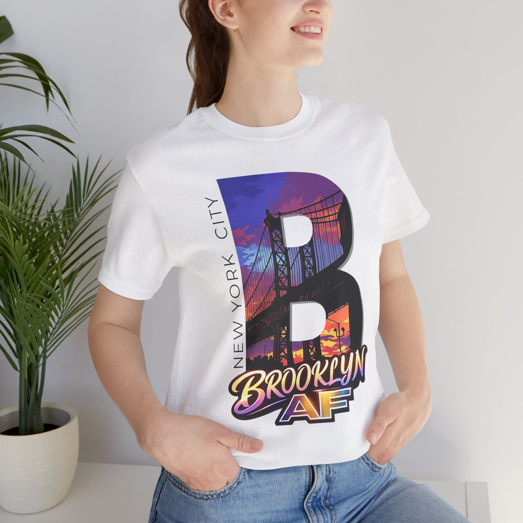 White tee worn by a female model near a plant, close-up of the "B" Brooklyn Bridge sunset skyline chest print.
