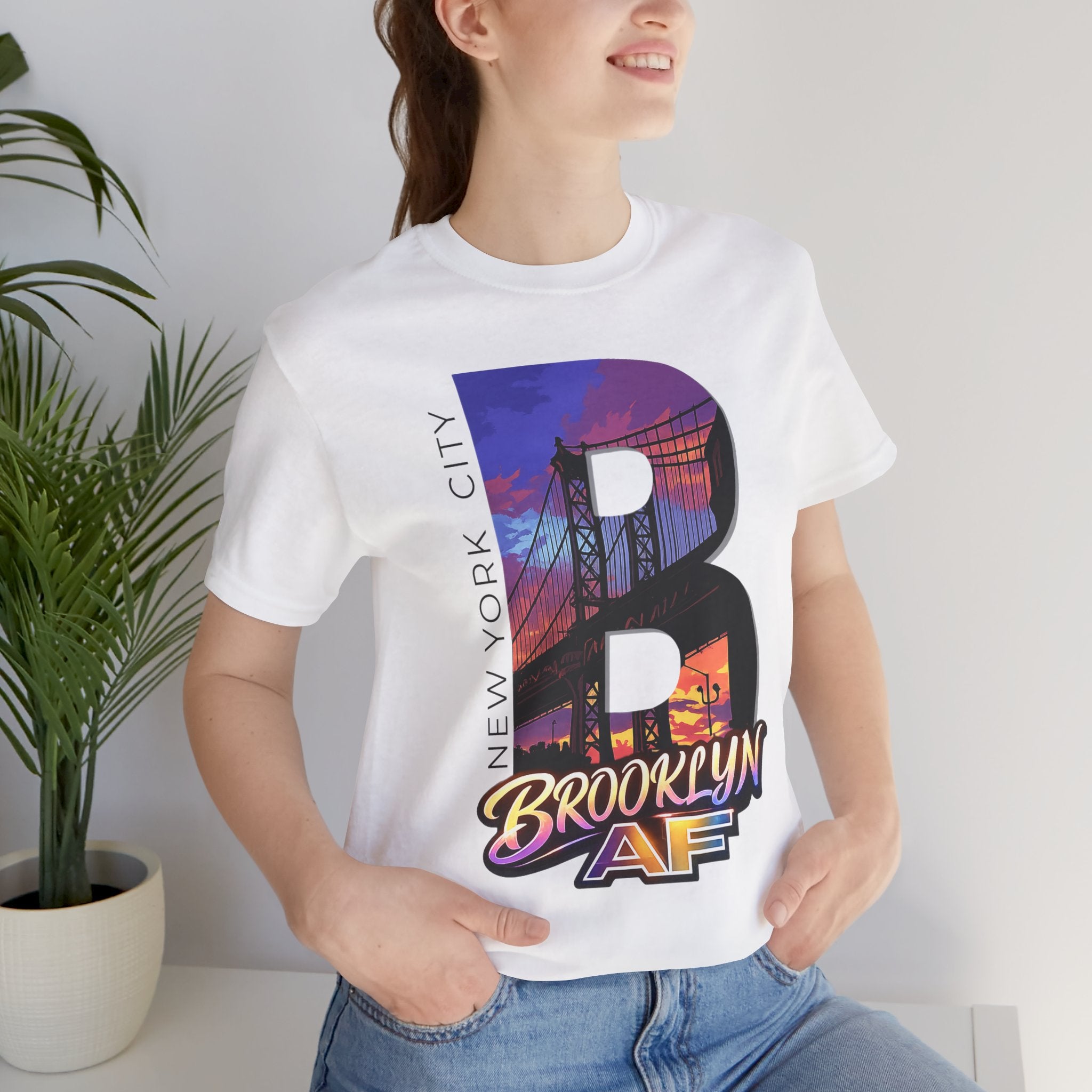 White tee worn by a female model near a plant, close-up of the "B" Brooklyn Bridge sunset skyline chest print.