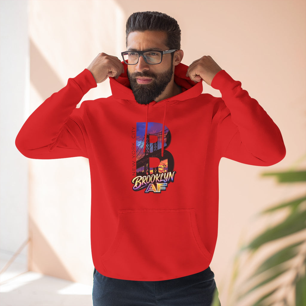 Red hoodie worn by a bearded male model pulling up the hood, bold B with a Brooklyn Bridge sunset skyline graphic.