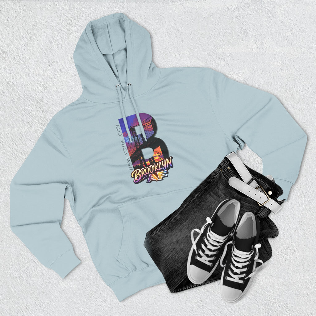 Pale blue hoodie styled with black jeans, belt, and sneakers, bold B featuring a Brooklyn Bridge sunset skyline graphic.