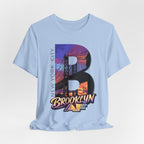 Light blue t-shirt flat lay, front view with full 'B' graphic, Brooklyn Bridge sunset skyline and 'Brooklyn AF' logo