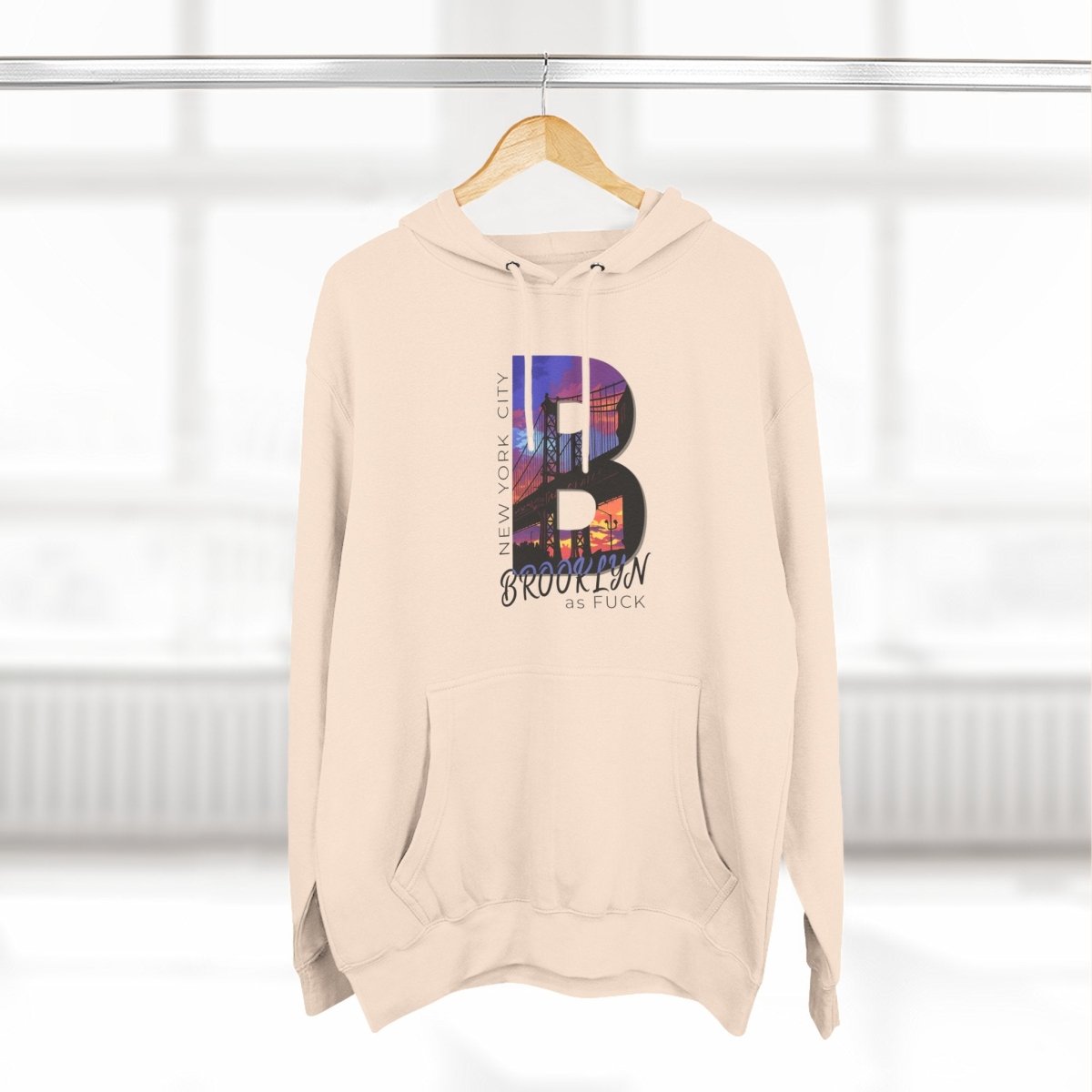Oatmeal Heather Brooklyn As Fuck hoodie on a wooden hanger by a window